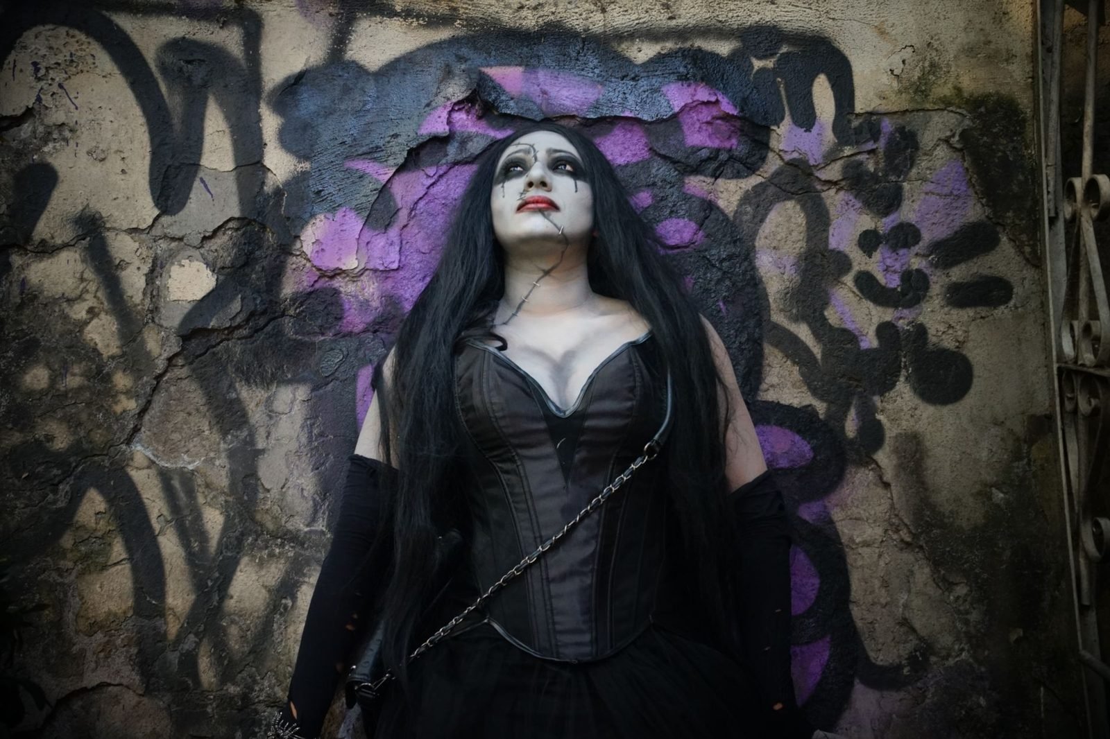 Mujer con corsé negro, maquillaje claro y vestuario gótico frente a un muro con grafitis morados